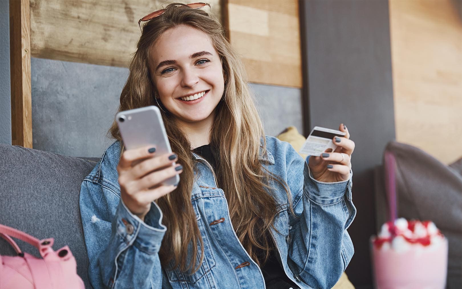 A smiling young woman in a denim jacket holding a smartphone and credit card while sitting on a couch, with shopping bags visible in the background.