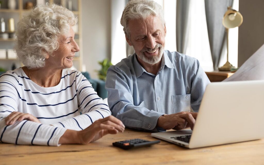 Senior couple smiling while using a laptop together at a wooden table in a modern home interior.