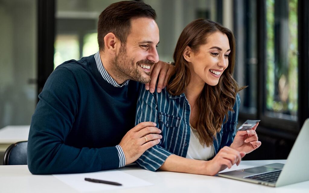 A smiling man and woman sit together at a desk looking at a credit card and laptop, appearing satisfied with an online purchase or financial transaction.