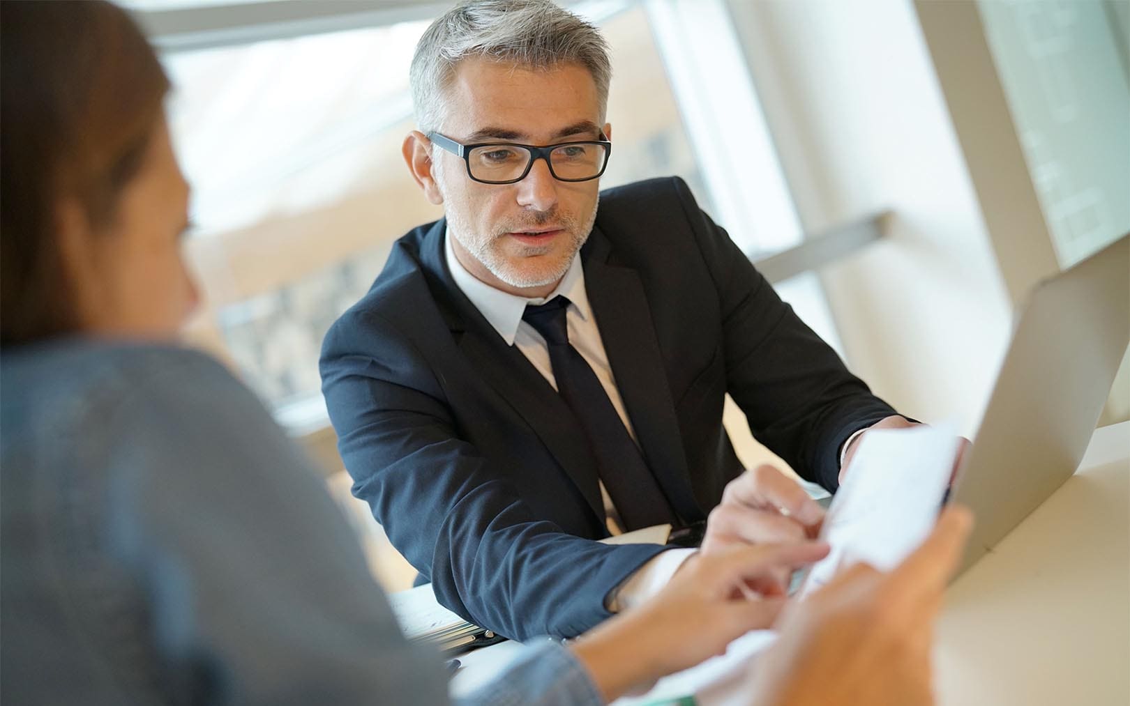 A mature businessman wearing glasses and a dark suit reviews a document with a female colleague in a bright, modern office setting.