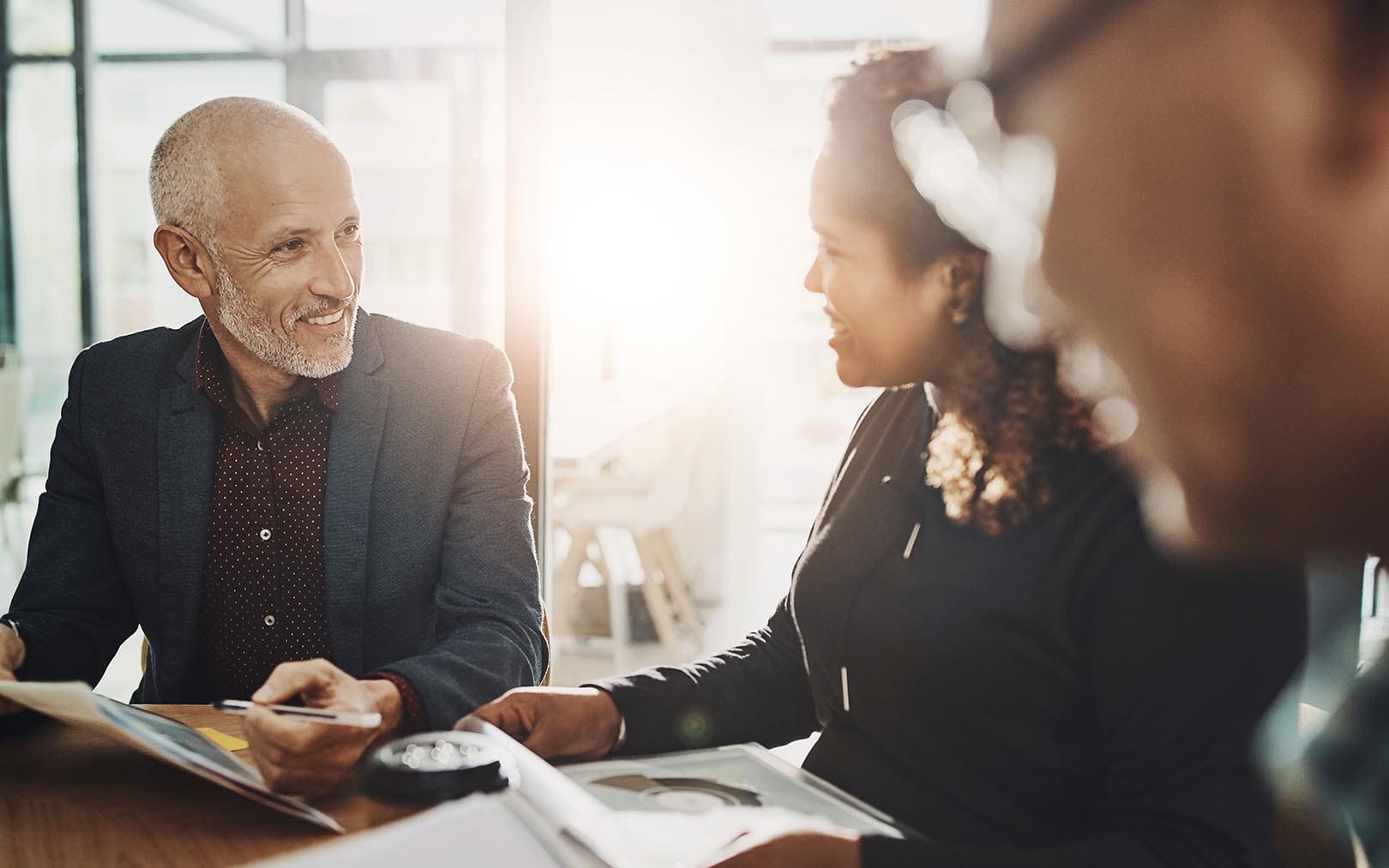 A diverse group of business professionals engaged in a collaborative meeting around a table with documents and materials, discussing ideas in a bright, modern office setting.