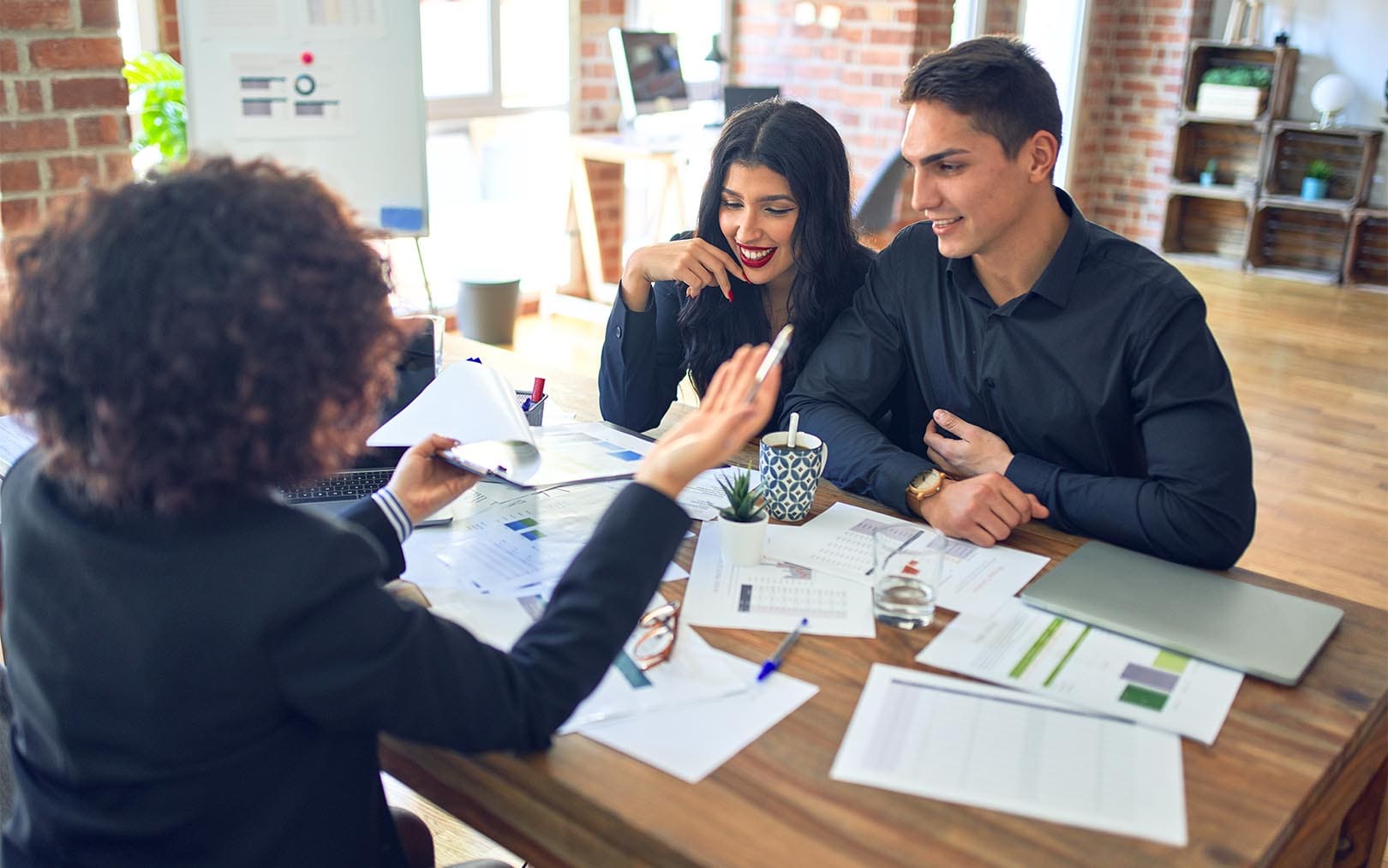 Three young professionals in business attire engaged in a collaborative meeting at a wooden table in a modern office space with exposed brick walls, reviewing documents and charts together.