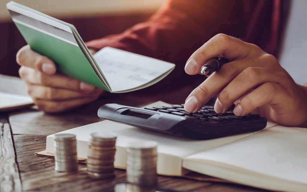 A person using a calculator and holding a green notebook while reviewing financial documents, with stacked coins visible on the desk, depicting budgeting and financial planning.