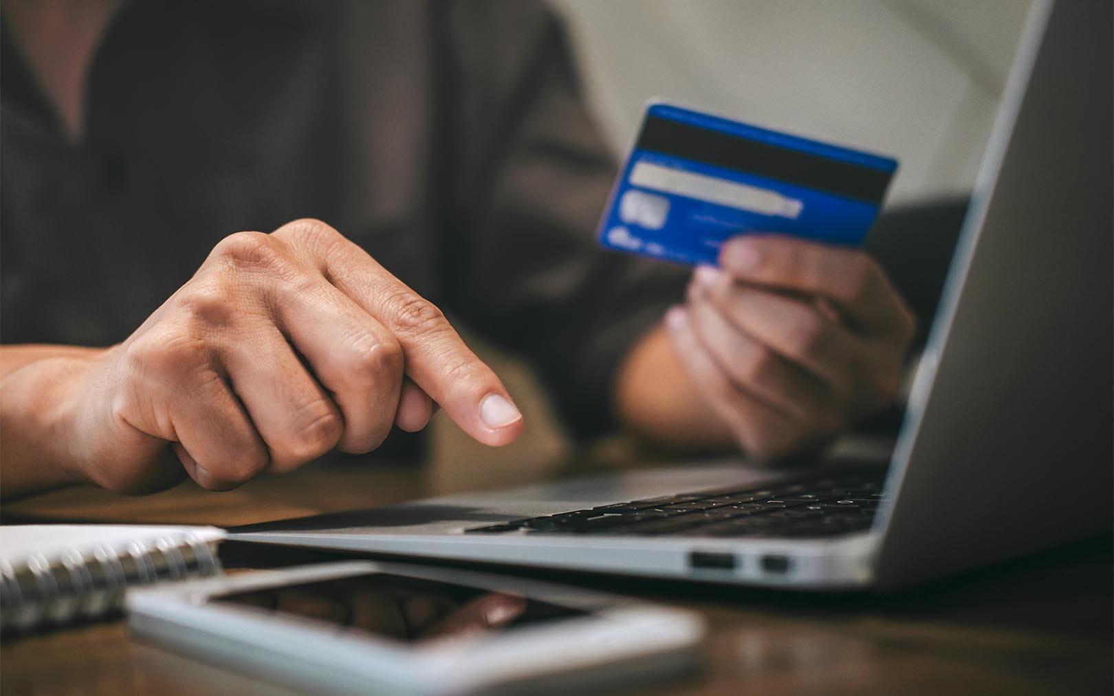 Person holding a blue credit card while using a laptop for online payment, with a smartphone on the desk.