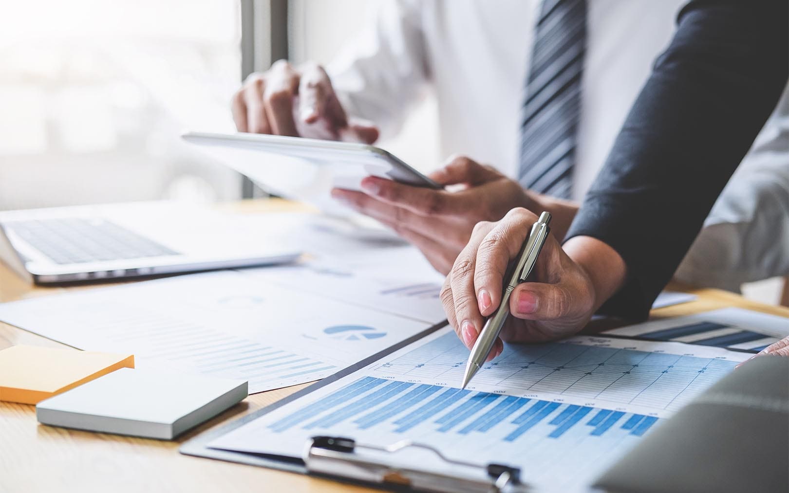 Business professionals reviewing financial charts and data on documents at a table with a laptop and tablet during a collaborative meeting.