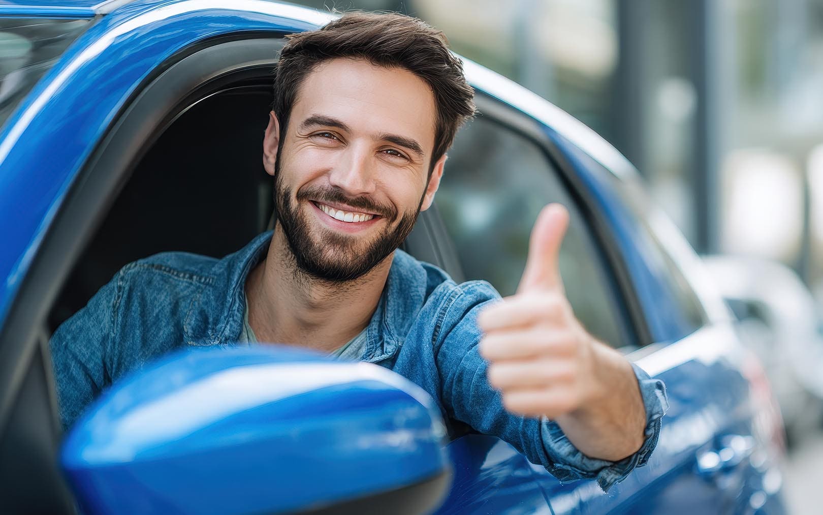 Happy man in denim shirt giving thumbs up while sitting in blue car with window open.