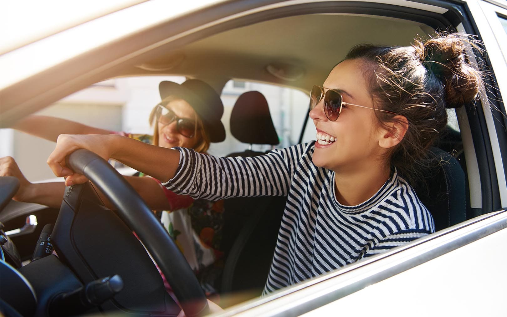 Two young women smiling while driving in a car on a sunny day, with the driver wearing sunglasses and a striped shirt holding the steering wheel.