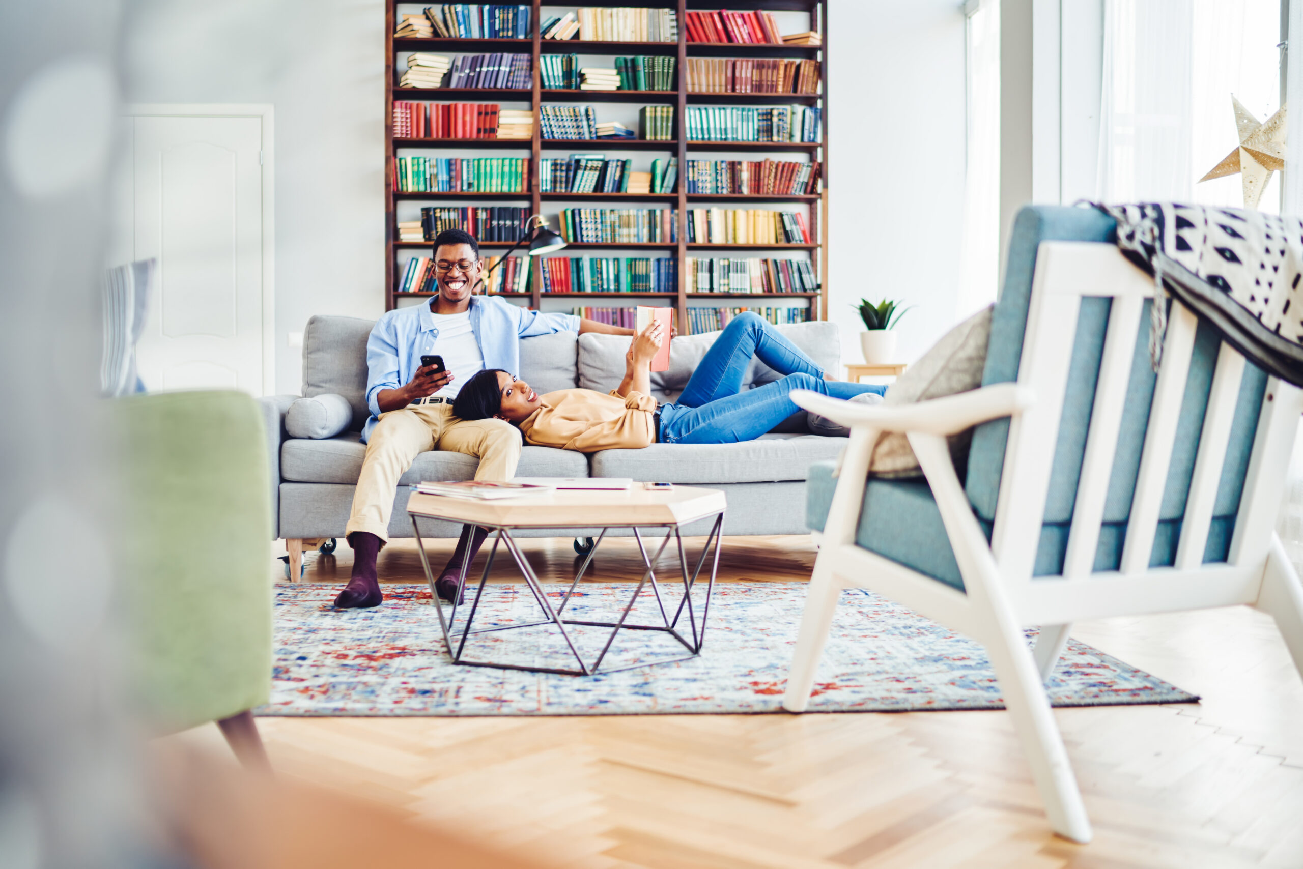 Happy relaxed young black woman with book lying on sofa next to cheerful husband with smartphone and together enjoying weekend at home