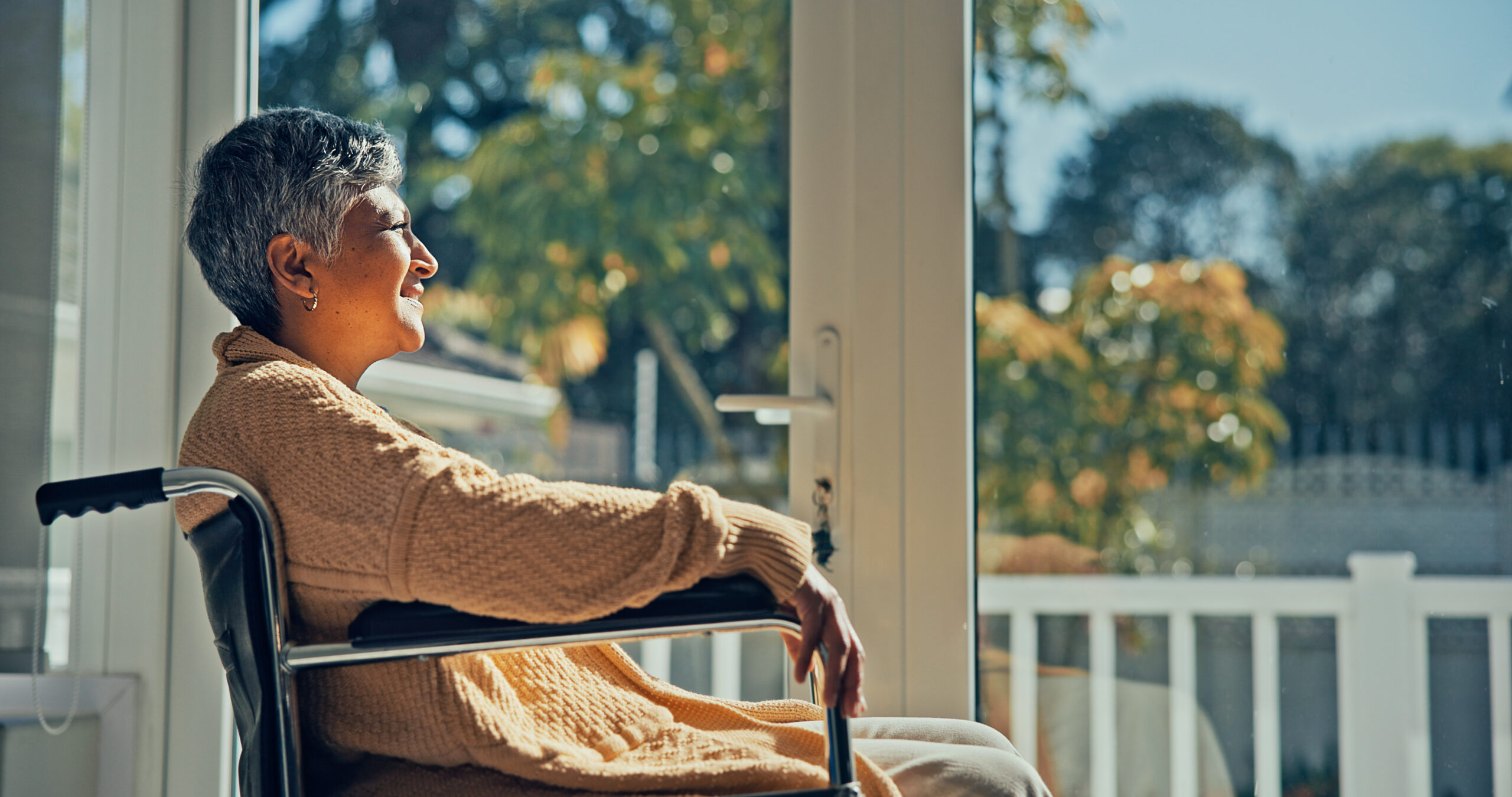 Mature woman sitting in wheelchair in nursing home looking out of window. Lonely disabled aged female patient in wheelchair looking through window in hospital corridor.