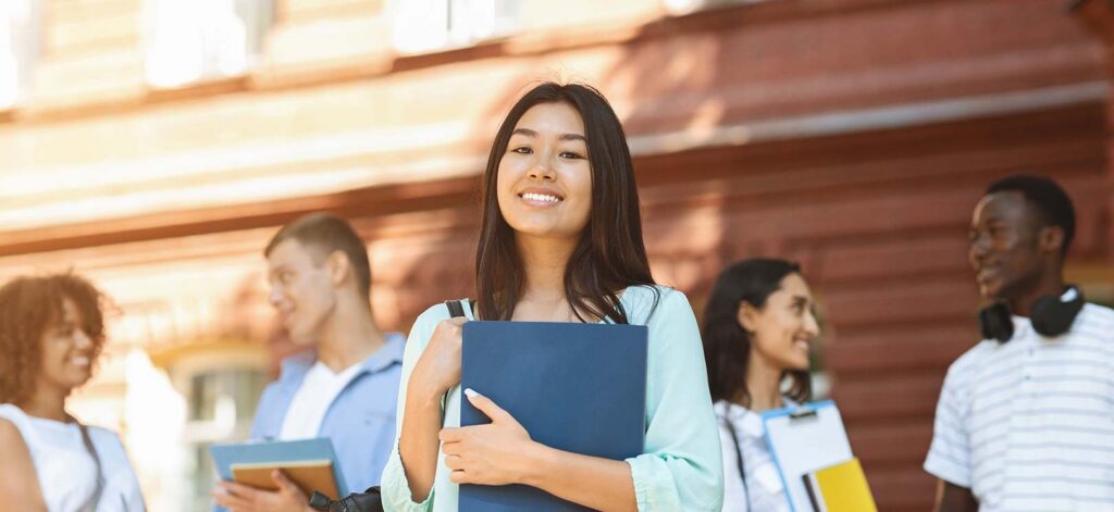 A smiling young Asian woman holding a blue notebook and backpack stands in focus in front of a brick building, with diverse students holding books blurred in the background.