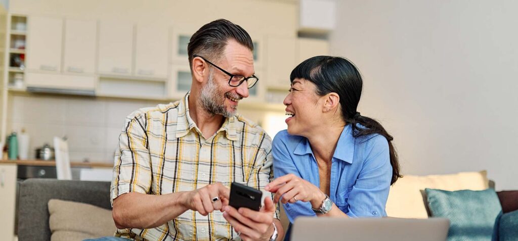 A man and woman sitting on a couch in a living room, smiling while looking at a smartphone together with a laptop and paperwork in front of them.