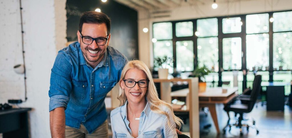 Two smiling professionals wearing glasses in a modern office workspace, looking at a laptop together at a desk with large windows in the background.