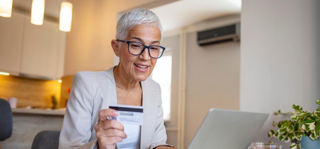 Smiling senior woman wearing glasses and beige cardigan holding a credit card while using a laptop at a desk in a modern kitchen.