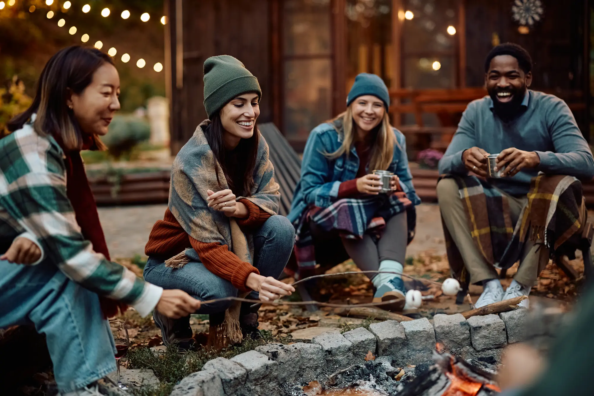 A diverse group of four young adults sitting around a fire pit in an urban courtyard at night, wearing winter clothing and holding drinks while roasting marshmallows. Warm string lights and building windows are visible in the background.